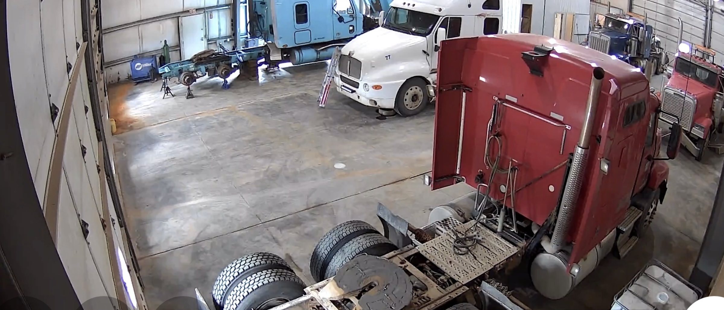 Inside the shop: a red semi tractor apart in the near bay with wheels and parts on the floor, and a second truck up in the bay behind it